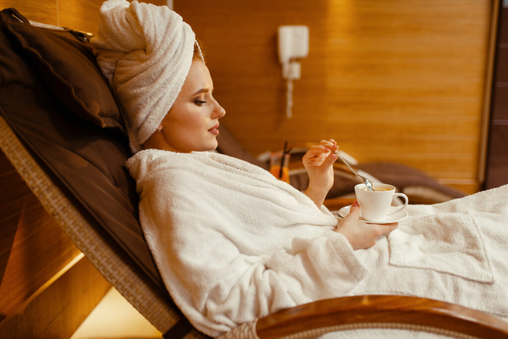 A sexy girl relaxing with a cup of coffee in a spa chair, surrounded by flowers and plants. She has a peaceful expression and is wearing a white bathrobe and flip flops. Luxury Treatments Aeonian Spa